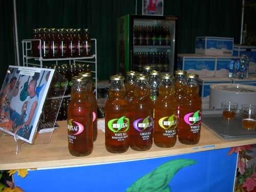 Rows of New Leaf bottled tea sit on a wooden counter, with a framed photo at left, a refrigerator and shelves of more bottles in the background.