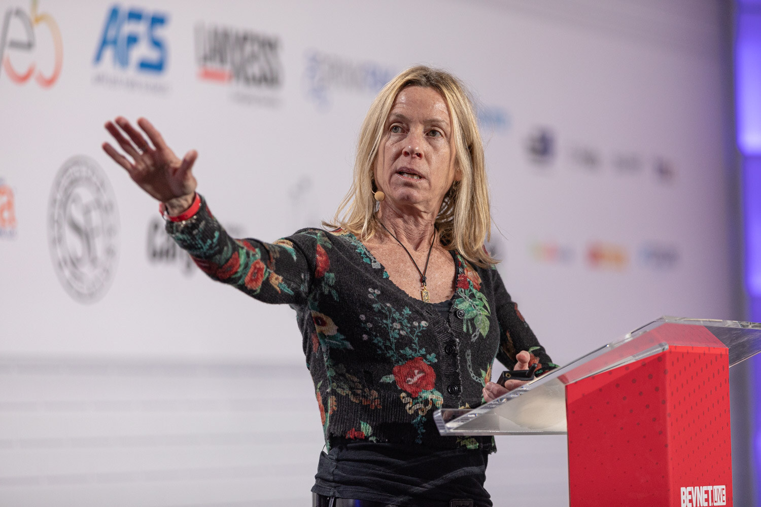 A woman with long blond hair in a floral cardigan extends one arm and speaks behind a red podium.