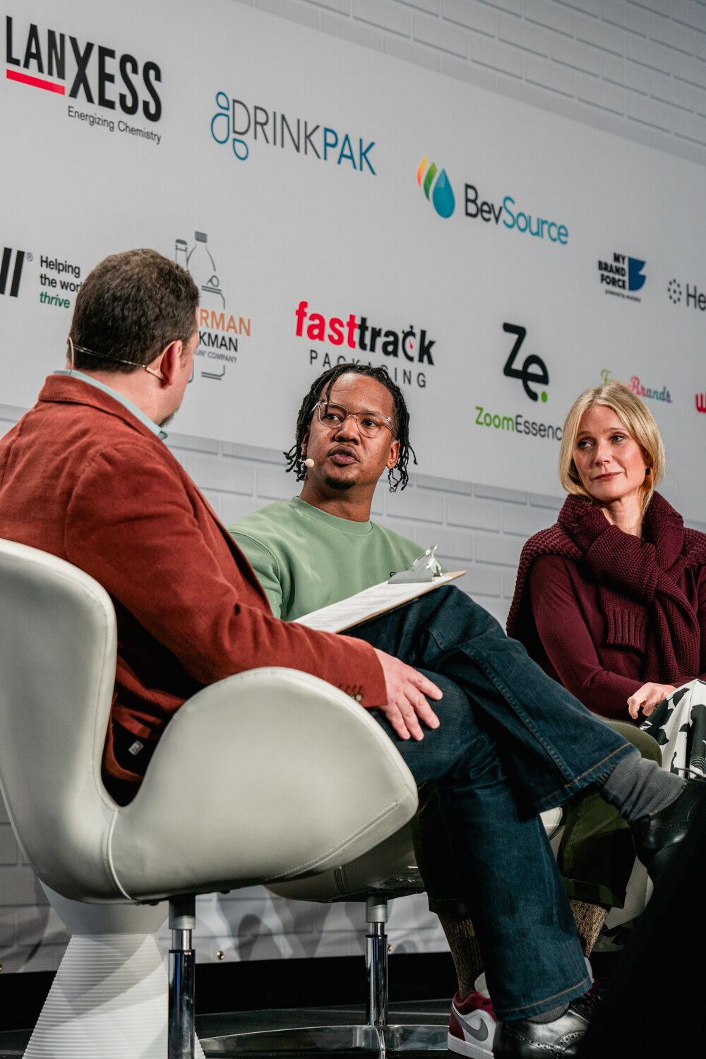 A vertical shot of a man seen from behind in a rust blazer, a man in a mint sweatshirt, and a woman in a maroon sweater seated in front of a sponsor wall.