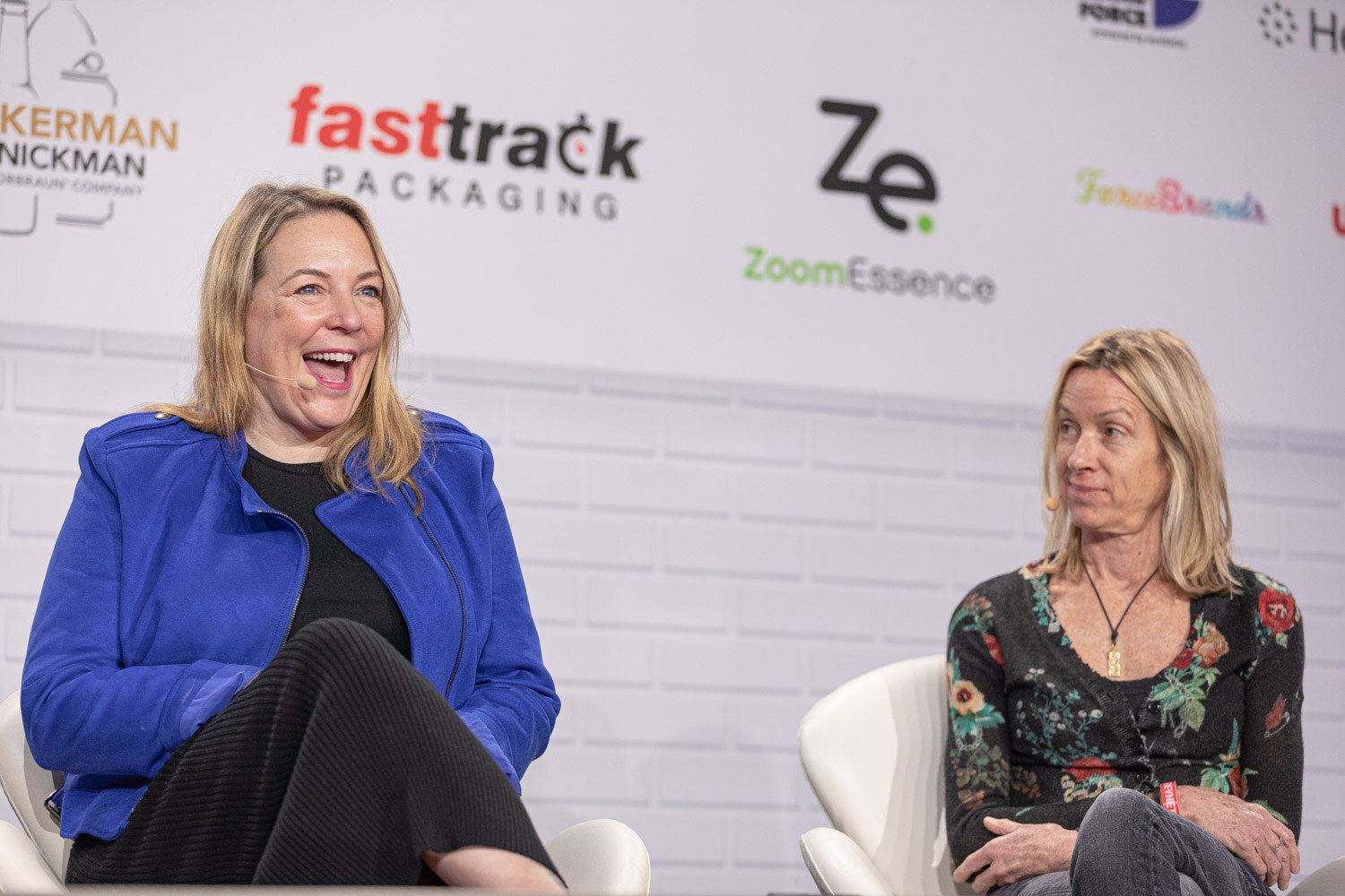 A woman in a bright blue jacket laughs while a woman in a floral top sits beside her in front of a sponsor wall with Fasttrack Packaging and ZoomEssence logos.