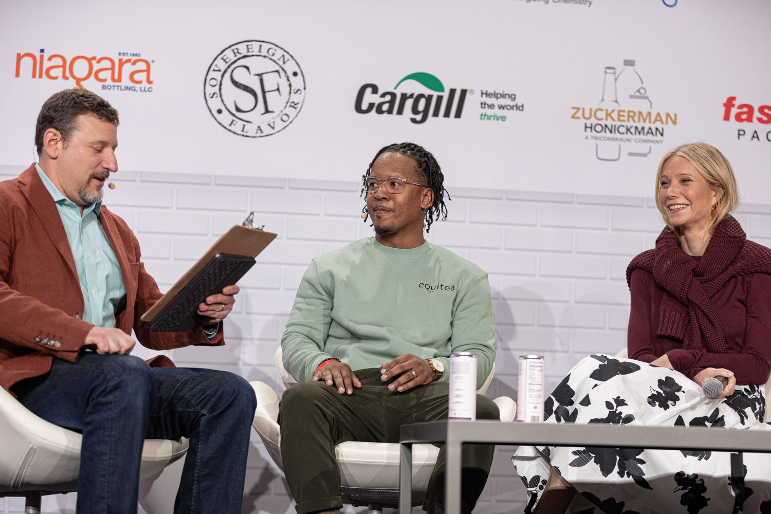 A man in a rust blazer holding a clipboard sits beside a man in a mint sweatshirt and a woman in a maroon sweater, in front of a wall of sponsor logos.
