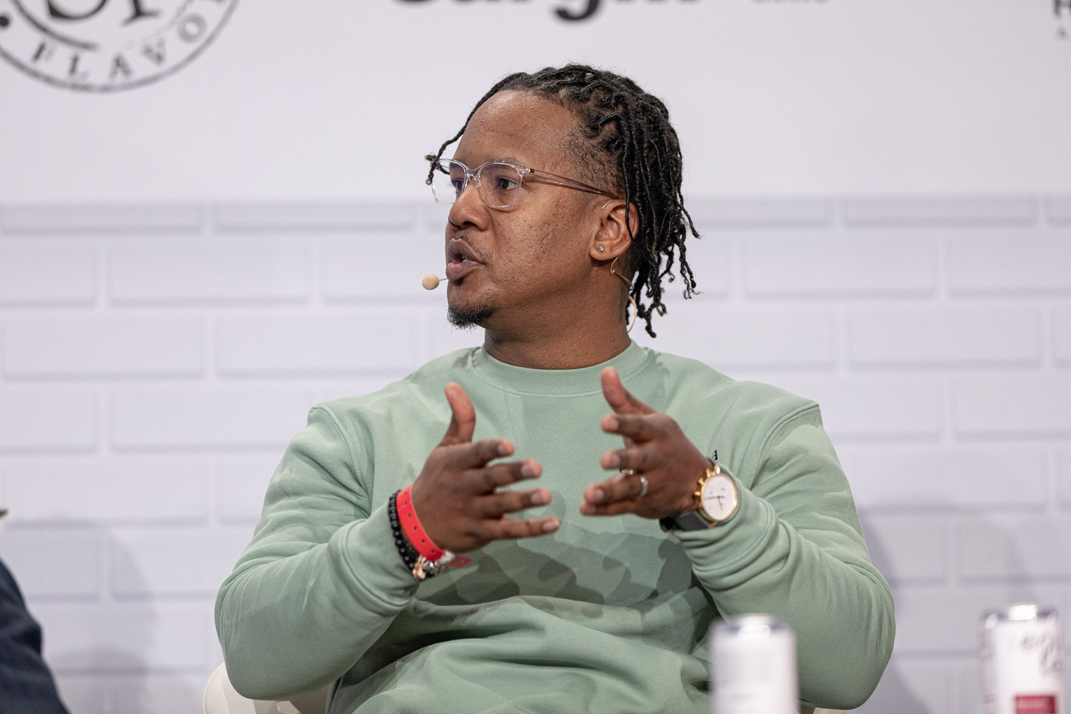 A man with twisted hair and clear-framed glasses in a mint sweatshirt with a headset microphone holds both hands open in front of him.