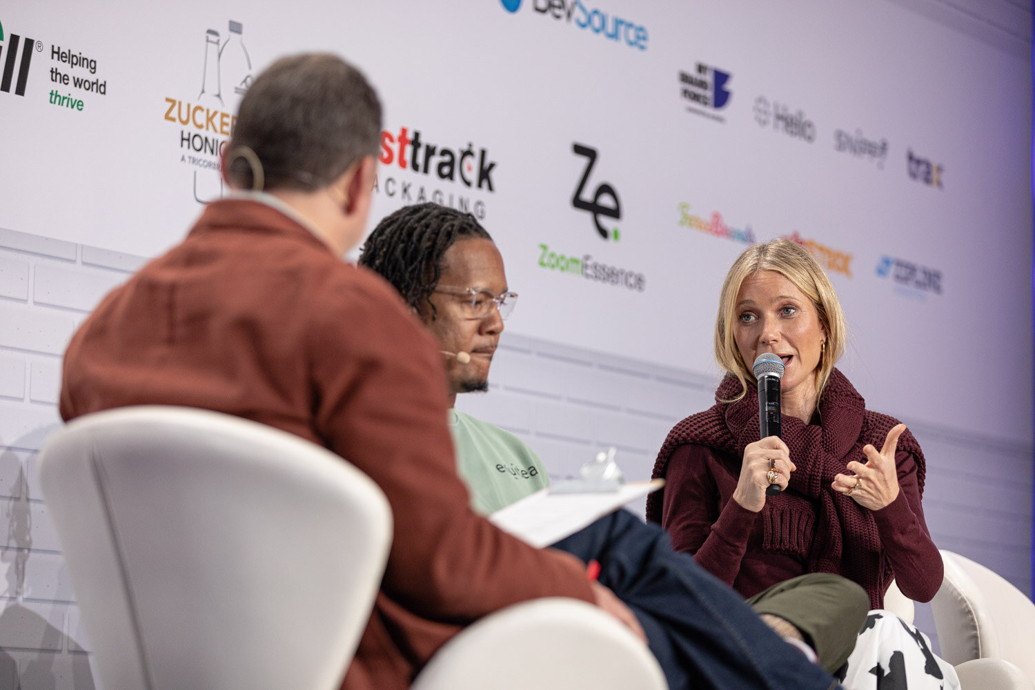 A woman in a maroon sweater holds a microphone and gestures while a man in a rust blazer, seen from behind, and a man in a mint sweatshirt listen.
