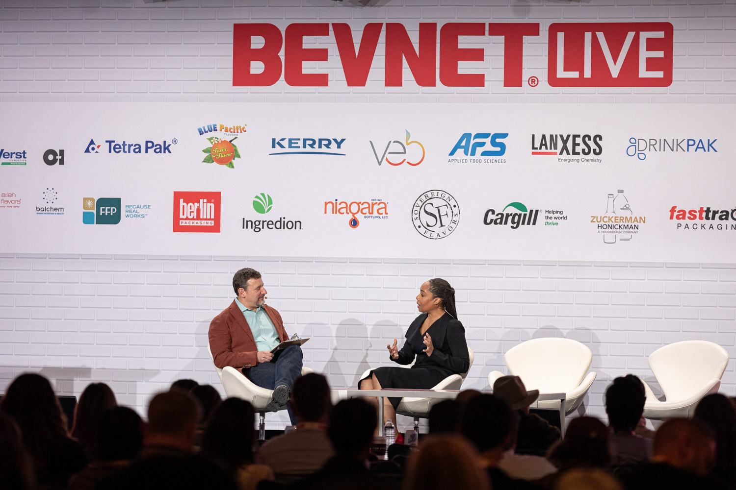 A wide shot of two people seated on white chairs on the main stage beneath a large BevNET Live sign and sponsor wall, with the audience's heads in the foreground.