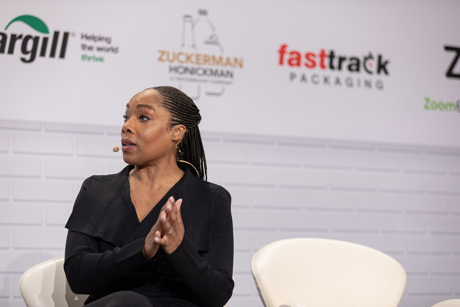 A woman with braided hair in a black wrap jacket and a headset microphone presses her palms together in front of a sponsor wall.