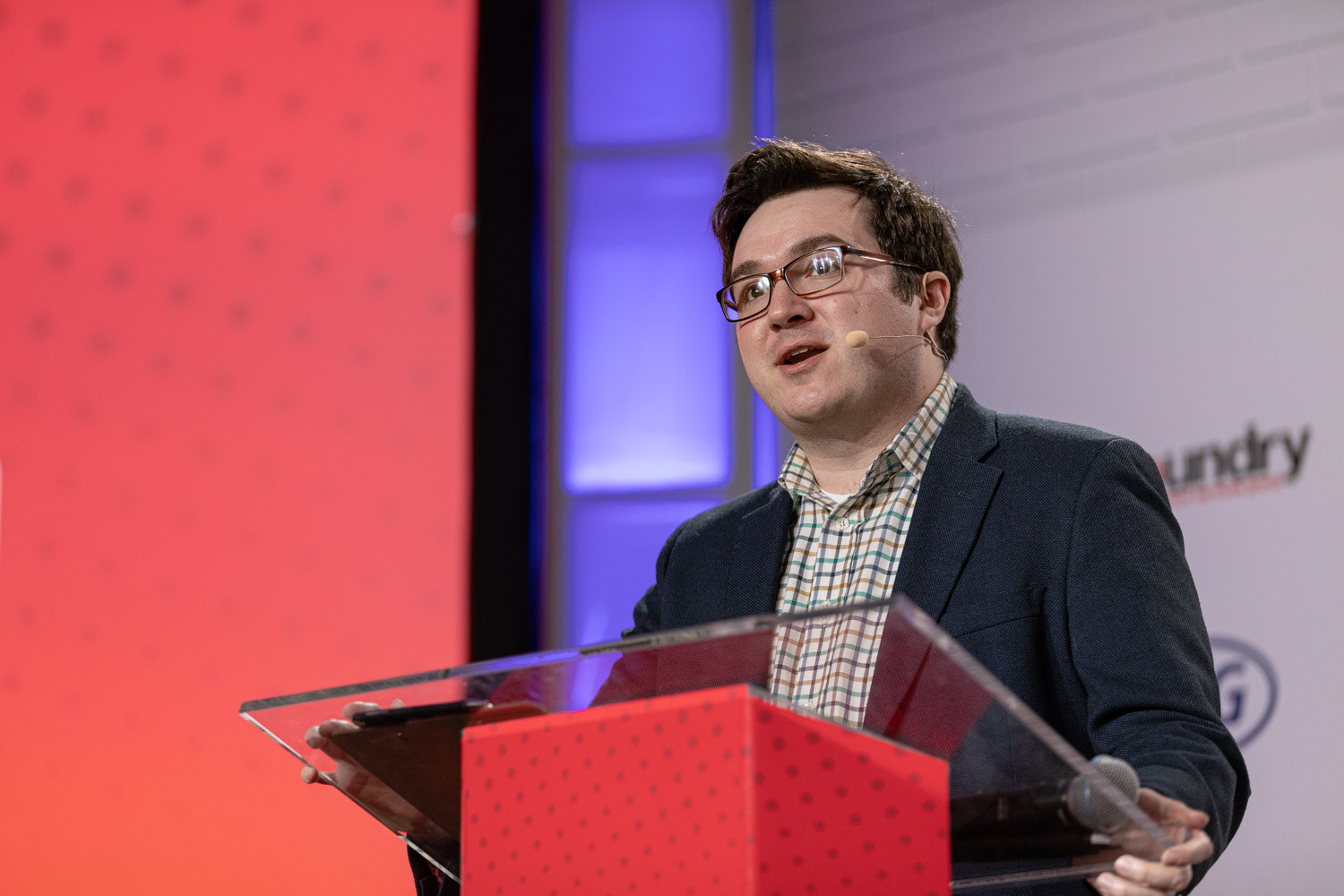 A man in glasses, a navy blazer and a checked shirt with a headset microphone speaks at a clear podium in front of an orange screen.