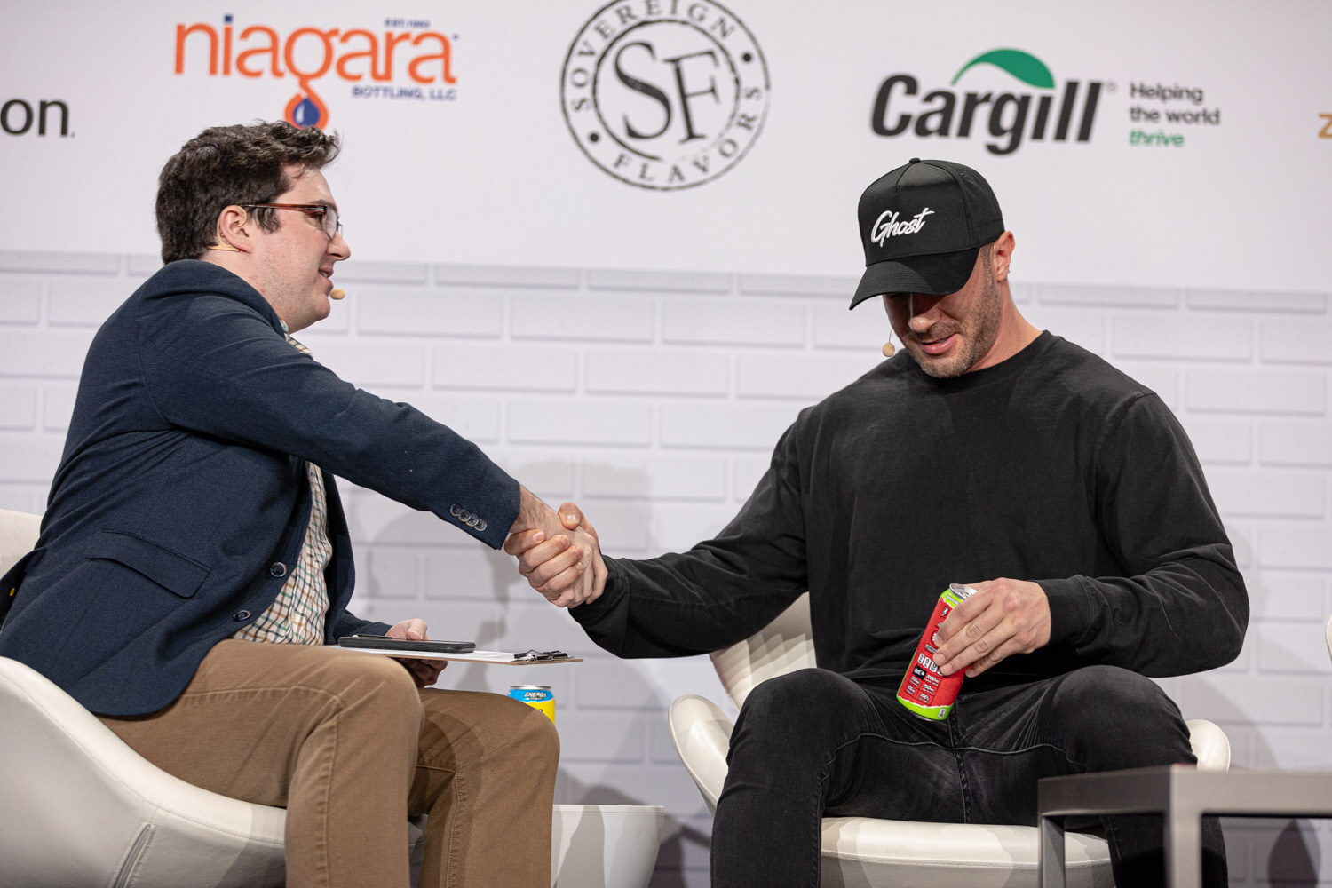 Two men seated in white chairs shake hands, one in a navy blazer and one in a black cap and black sweatshirt holding a red can.