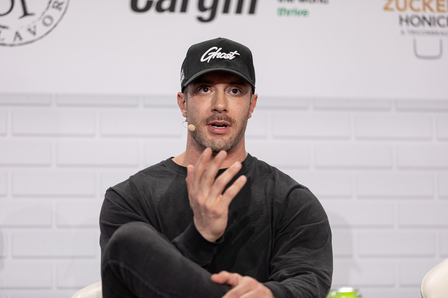 A man in a black Ghost baseball cap and black sweater with a headset microphone gestures with one hand in front of a white brick wall.