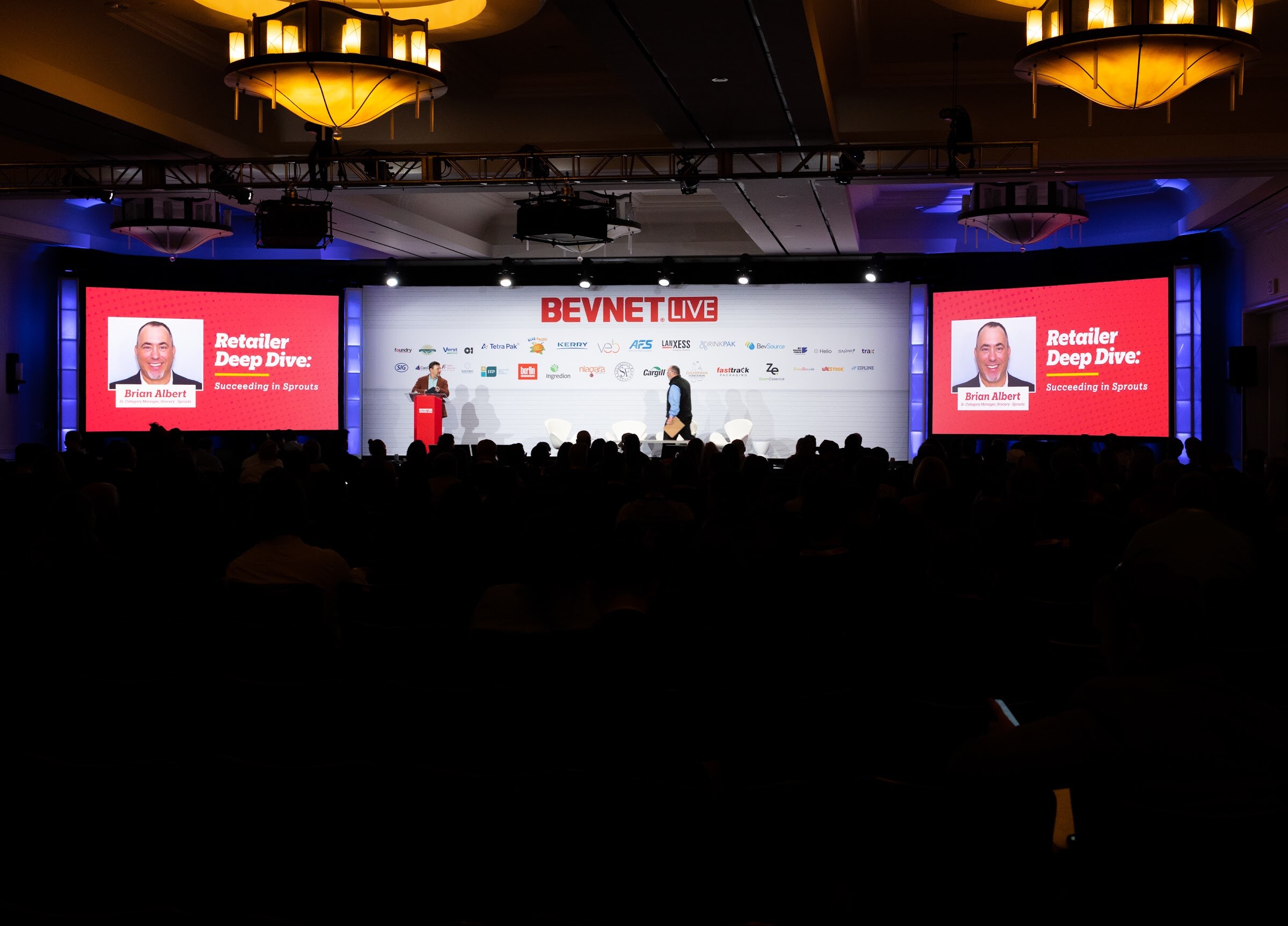A dark ballroom with a stage in the center and two red screens reading Retailer Deep Dive: Succeeding in Sprouts.