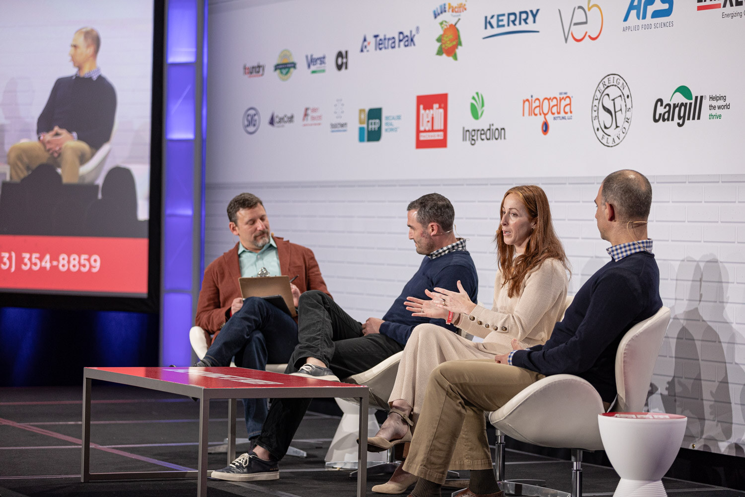 Four people on white chairs on a stage, with a large video screen at left showing one of the panelists.