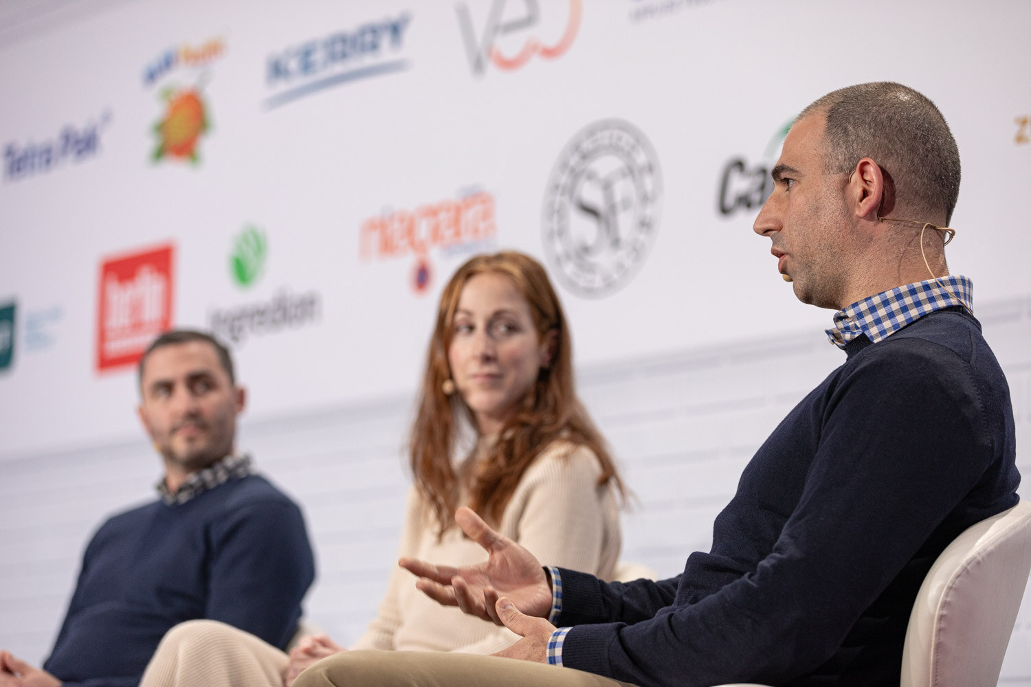 A bald man in a navy sweater and checked collar gestures in the foreground while a woman and another man listen behind him.