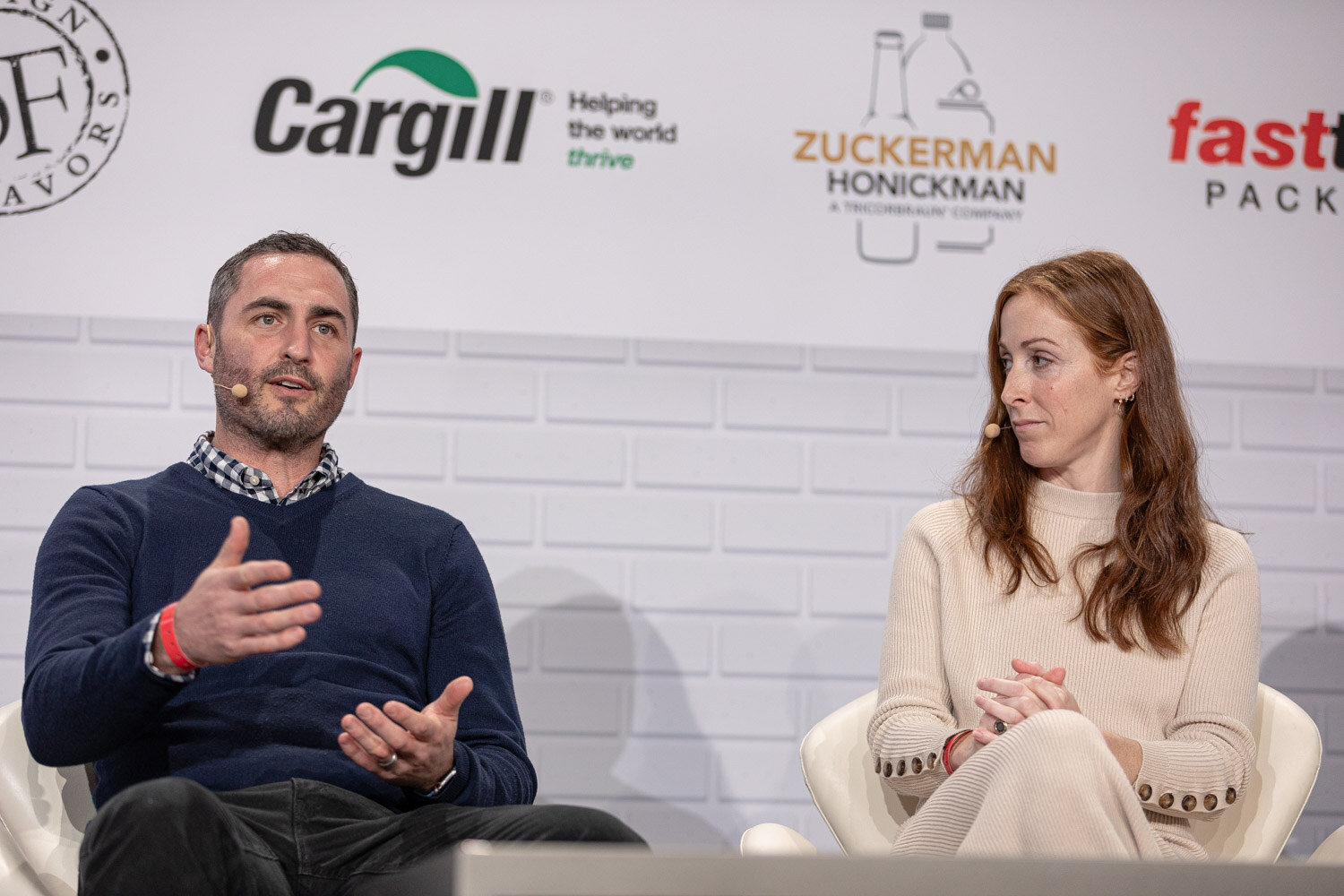 A man in a navy sweater with a headset microphone gestures while a woman with long red hair in a cream sweater listens.