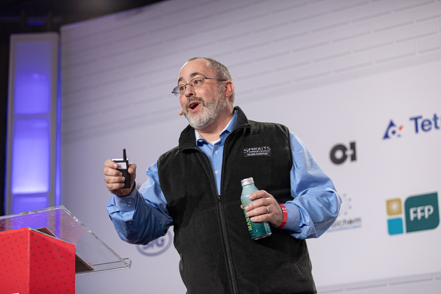 A bearded man in glasses and a black fleece vest labeled Sprouts holds a clicker in one hand and a green bottle in the other.