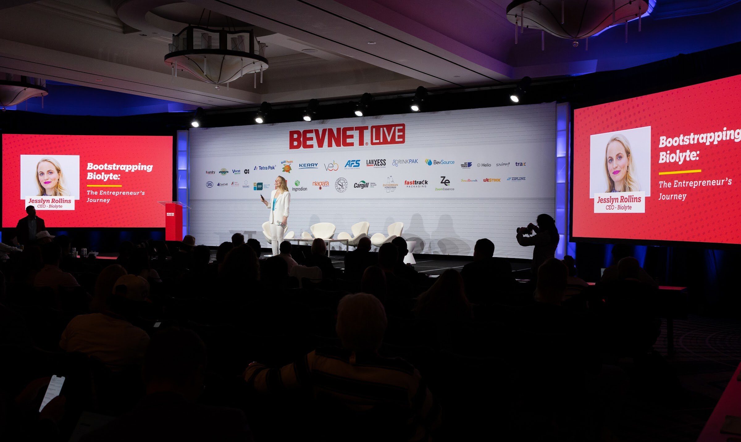 A dark ballroom with a stage in the center and two red screens reading Bootstrapping Biolyte: The Entrepreneur's Journey.