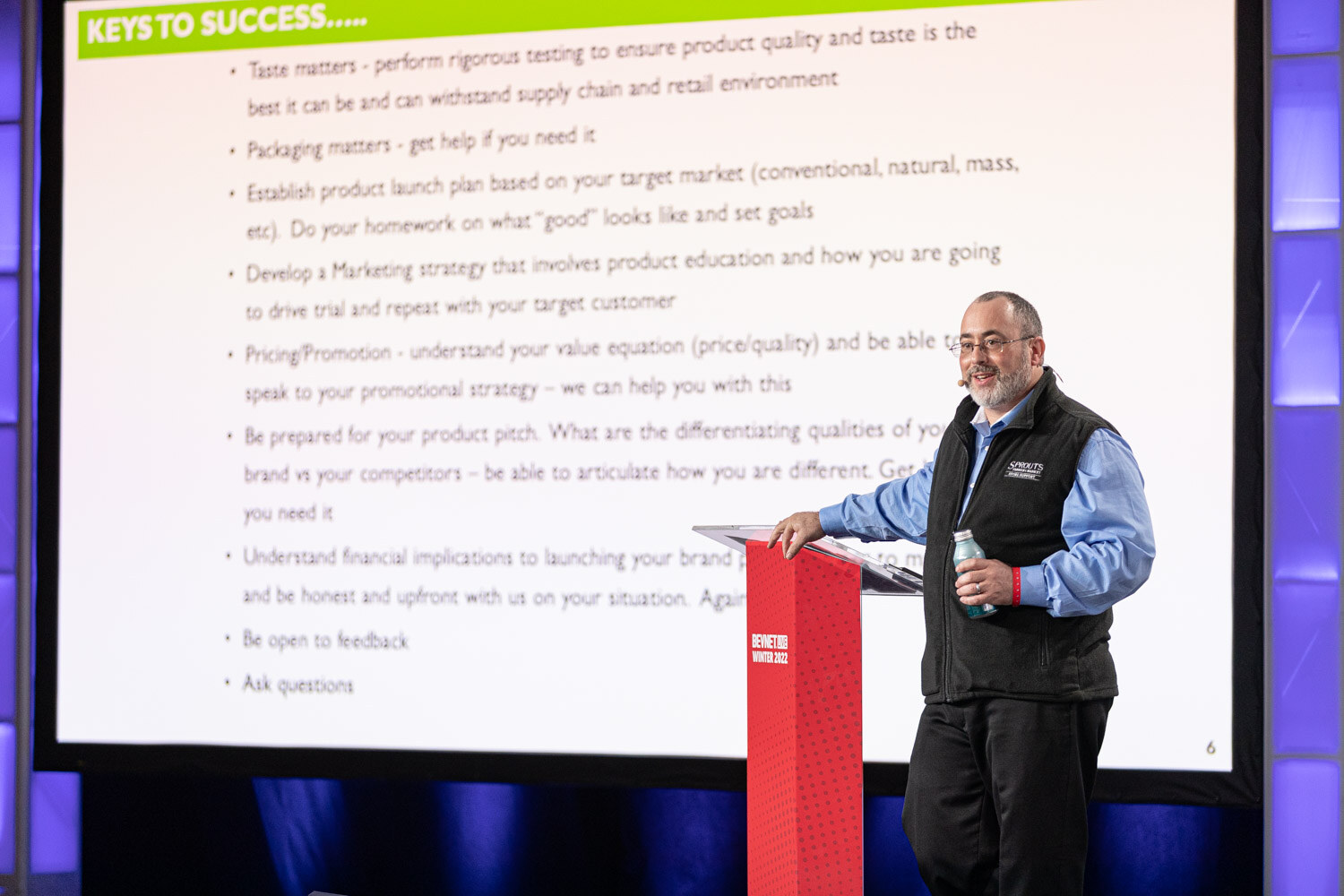 A man in a black Sprouts vest stands at a red podium next to a large screen showing a list titled Keys to Success.