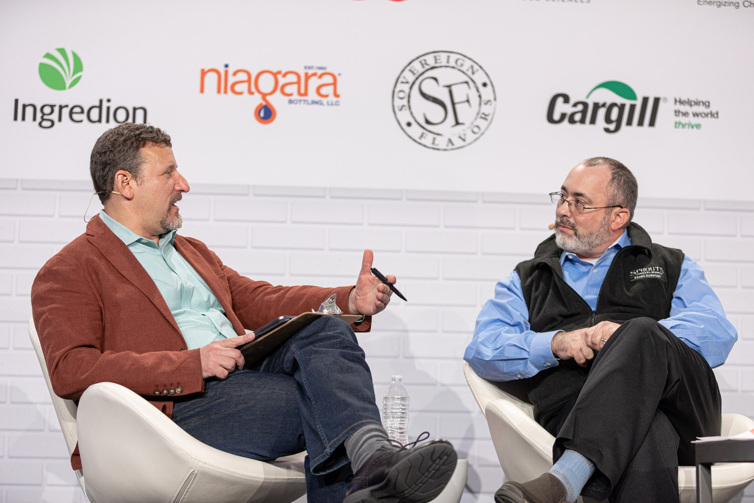 A man in a rust blazer holding a pen and a bearded man in a Sprouts vest sit across from each other in front of a sponsor wall.