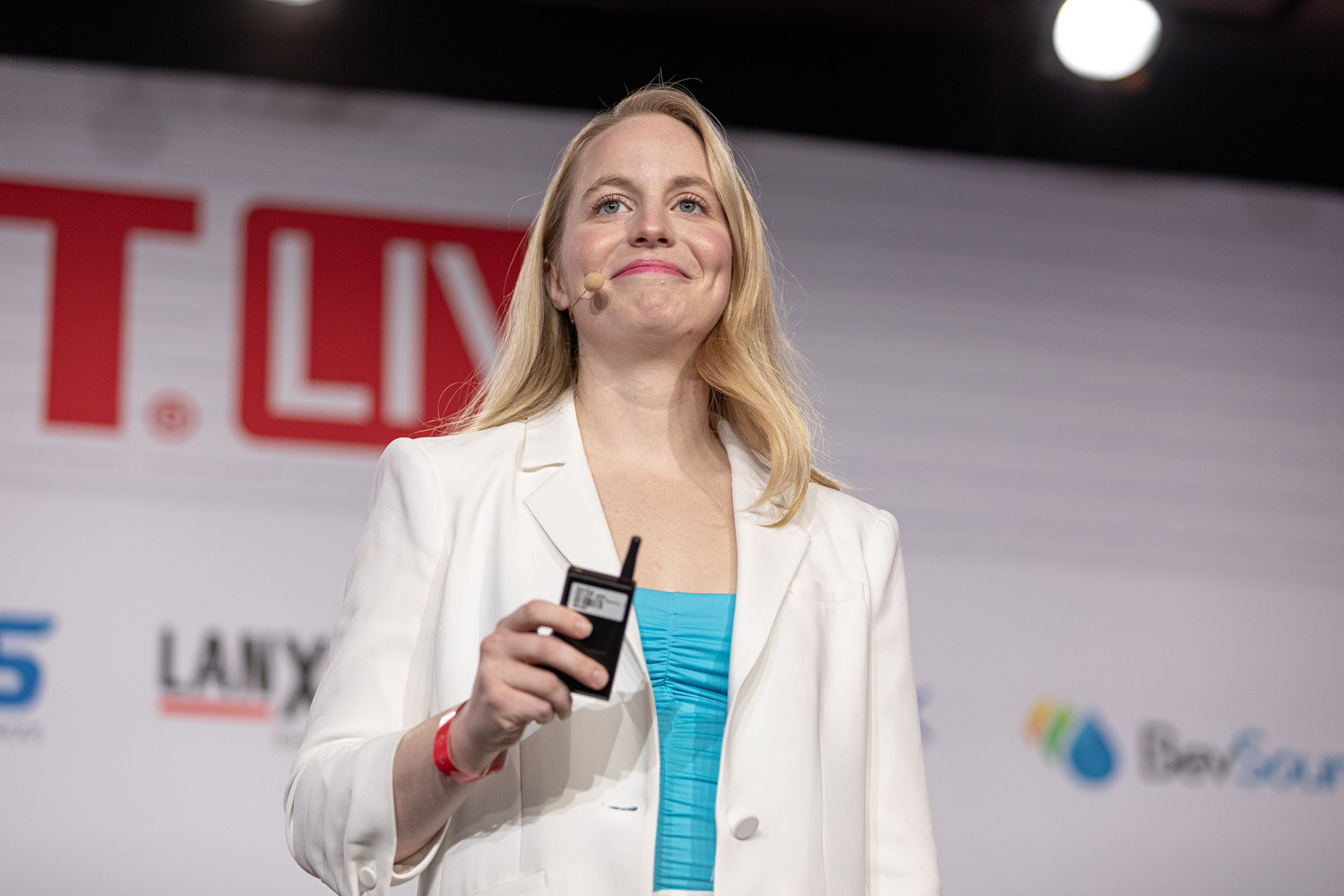 A blond woman in a white blazer and turquoise top holds a clicker and smiles in front of a BevNET Live sign and sponsor wall.