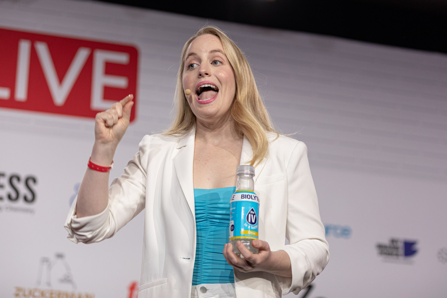 A blond woman in a white blazer and turquoise top raises a fist and holds a Biolyte bottle in the other hand while speaking.