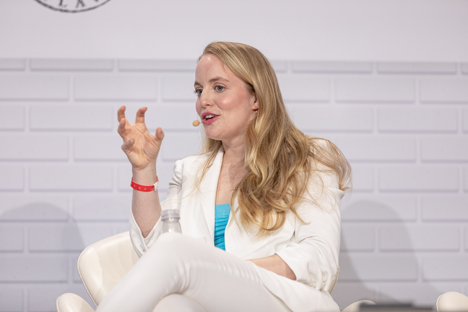 A blond woman in a white blazer sits with her legs crossed, one hand raised, in front of a white brick backdrop.