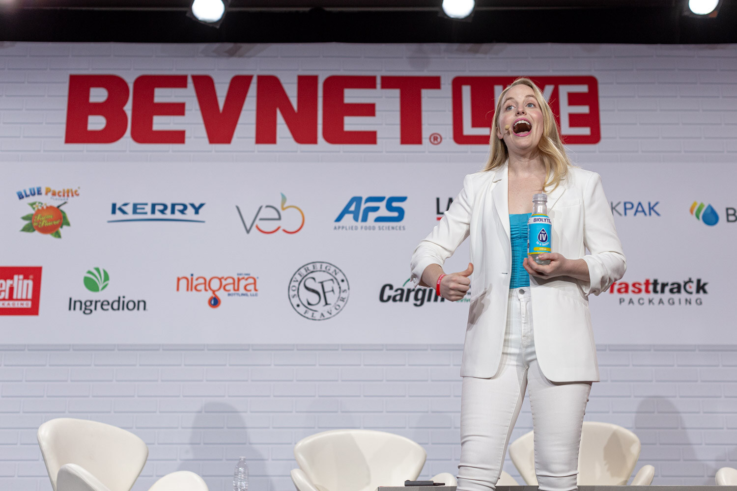 A woman in white with a Biolyte bottle stands center stage in front of the BevNET Live sponsor wall with her mouth open in mid-sentence.