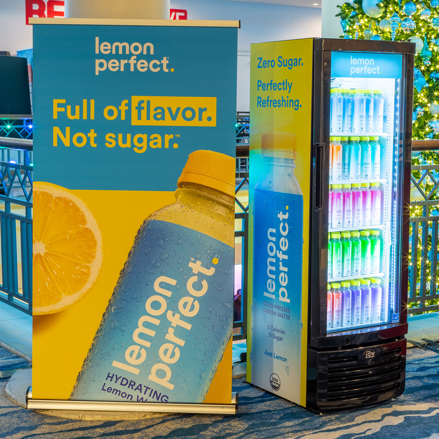 A yellow and blue Lemon Perfect banner reading Full of flavor, Not sugar beside a lit cooler of lemon water bottles and a holiday tree.