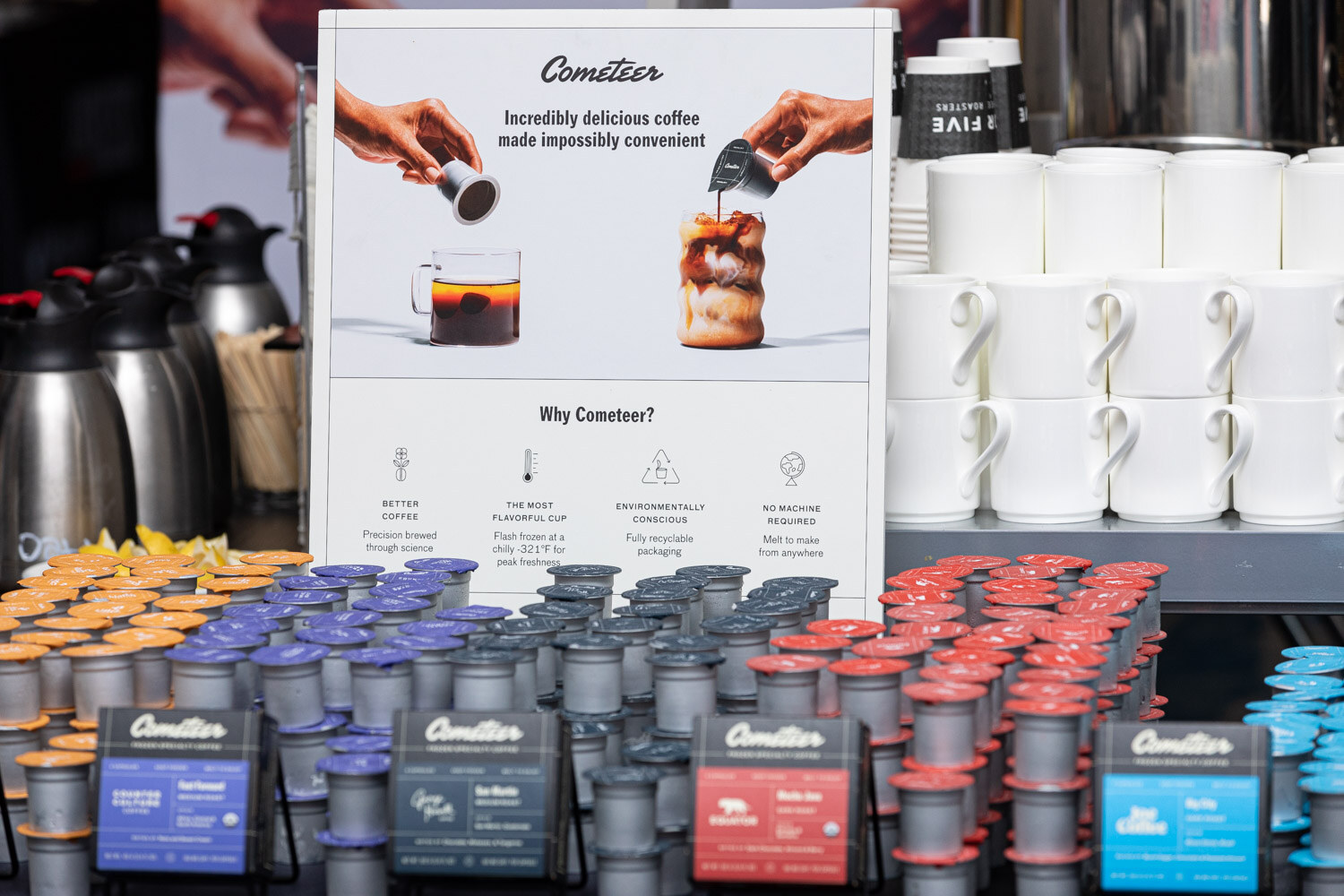 A table covered with rows of silver Cometeer coffee pods in orange, blue, red and light blue next to white mugs, thermal carafes and a sign reading Incredibly delicious coffee made impossibly convenient.