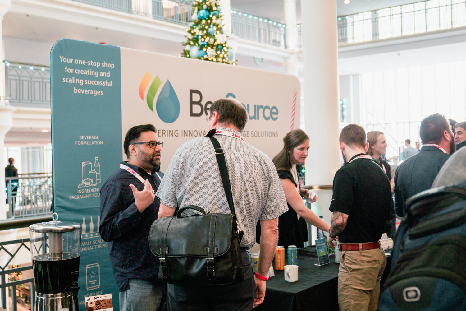 Attendees gather around a table in front of a BevSource banner in a bright atrium with a decorated Christmas tree.