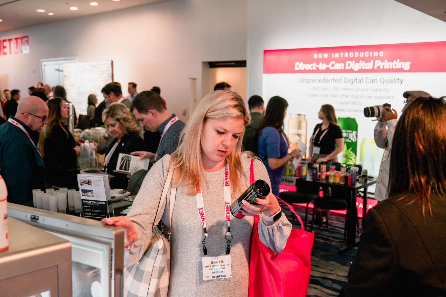 A blond woman with a red bag holds a can and reads its label at an exhibitor table, with a sign in the background reading Direct-to-Can Digital Printing.