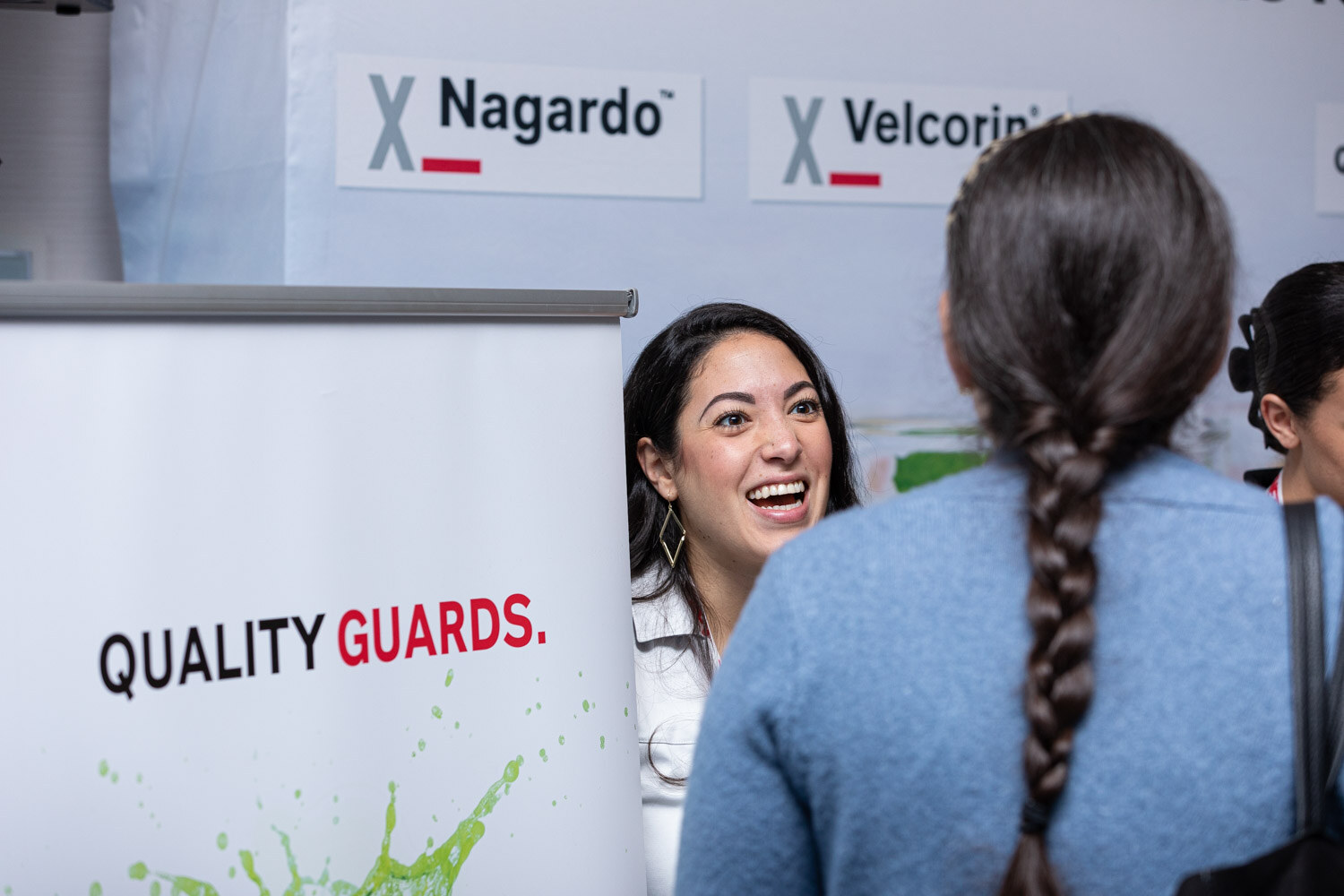 A smiling woman peeks out from behind a banner reading Quality Guards with Nagardo and Velcorin on the wall behind, while an attendee with a braid faces her.