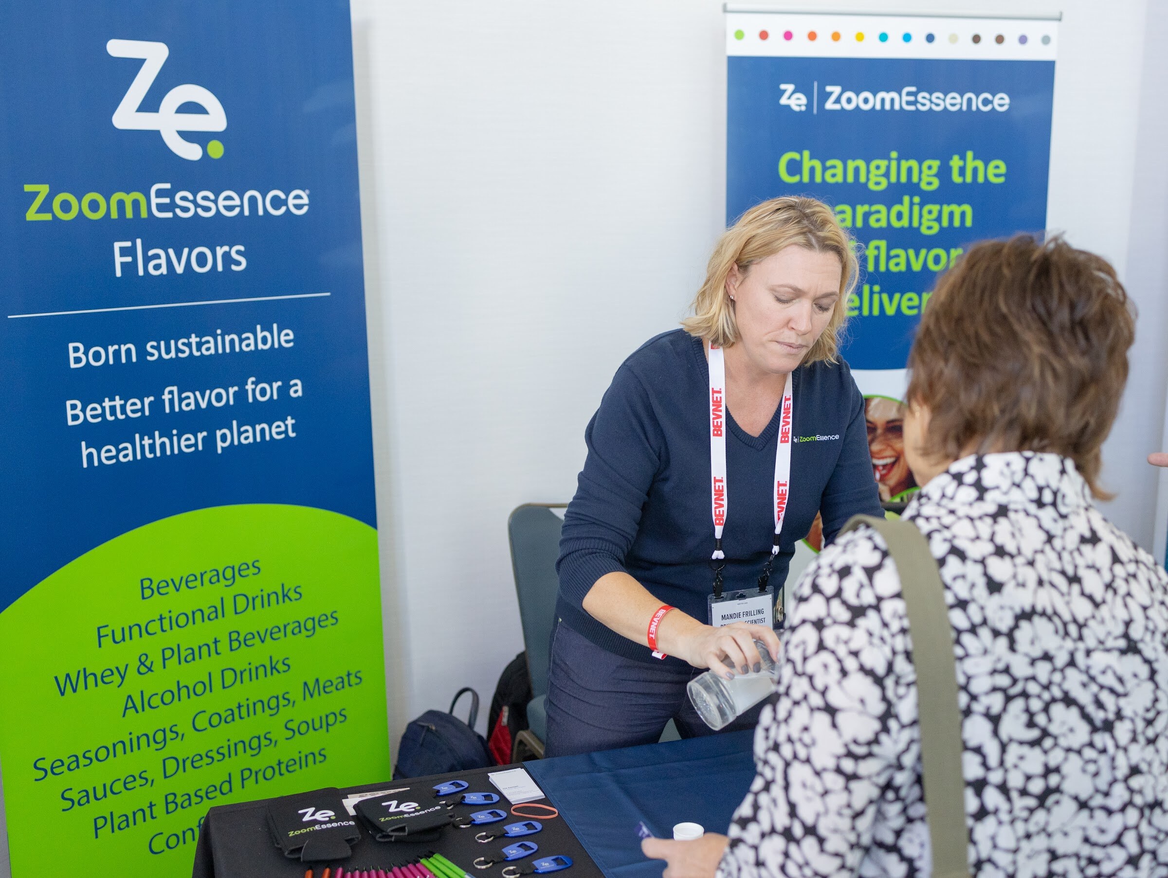 A blond woman in a navy shirt with a BevNET lanyard speaks with an attendee in a patterned top in front of a ZoomEssence Flavors banner.