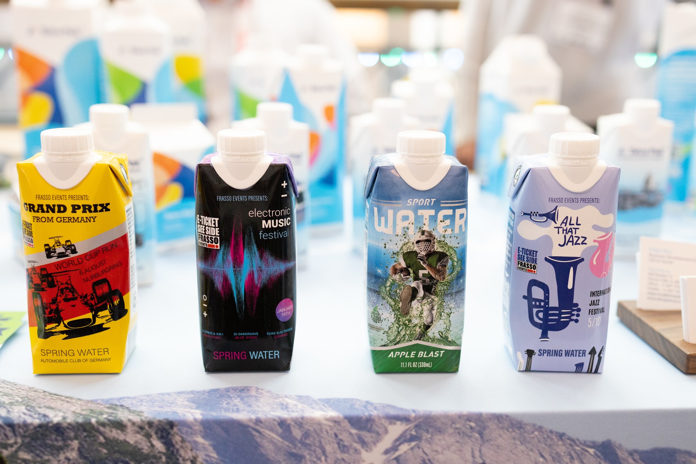 Four cartons of spring water and apple blast drinks with graphic labels, including All That Jazz and Grand Prix, sit on a white table.