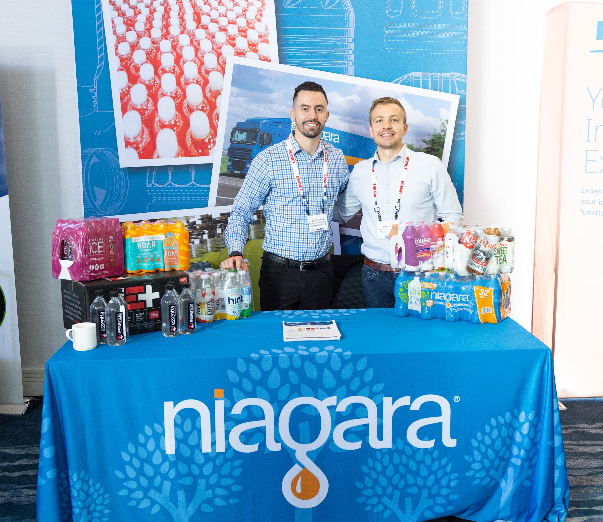 Two men in blue checked shirts with BevNET lanyards stand behind a blue table covered with a Niagara cloth and packs of bottled water.