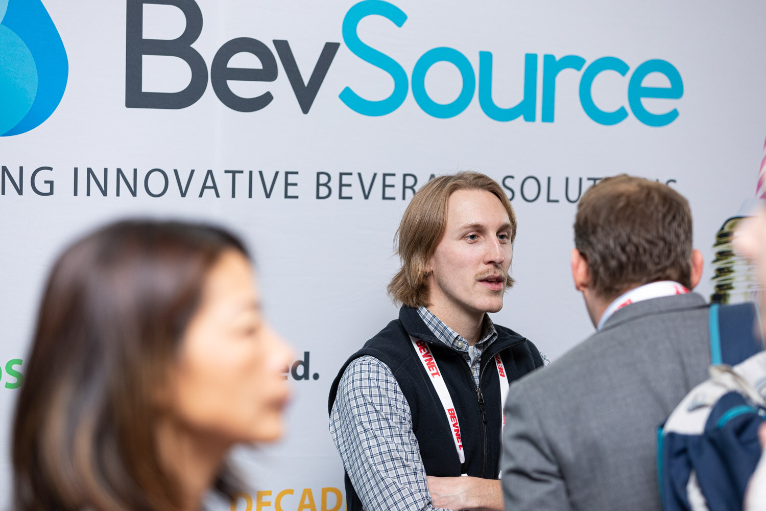 A man in a vest and a checked shirt with a BevNET lanyard talks with others in front of a large BevSource banner reading Delivering Innovative Beverage Solutions.