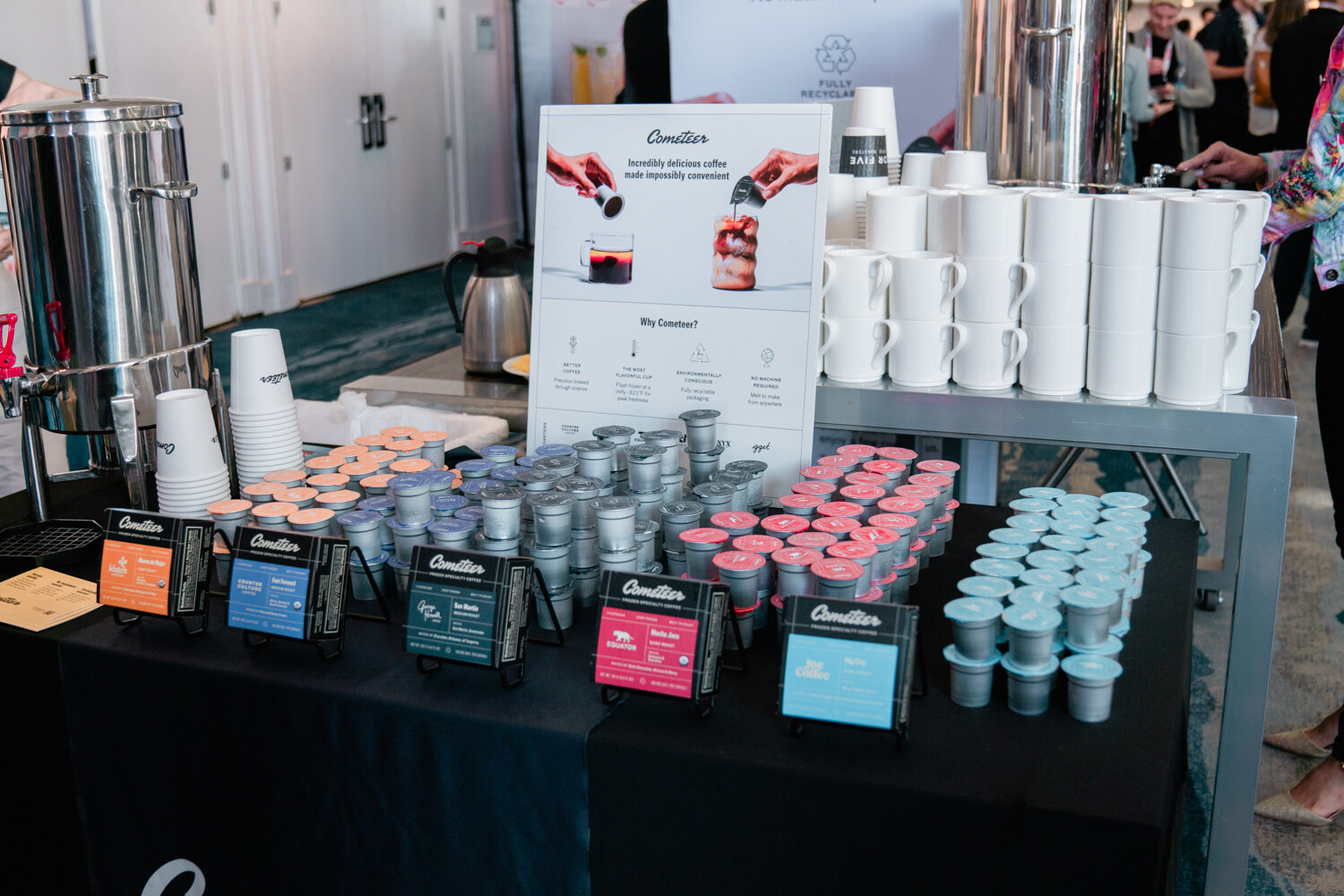 A table with rows of Cometeer pods in several colors, a large urn and stacks of white mugs and cups.