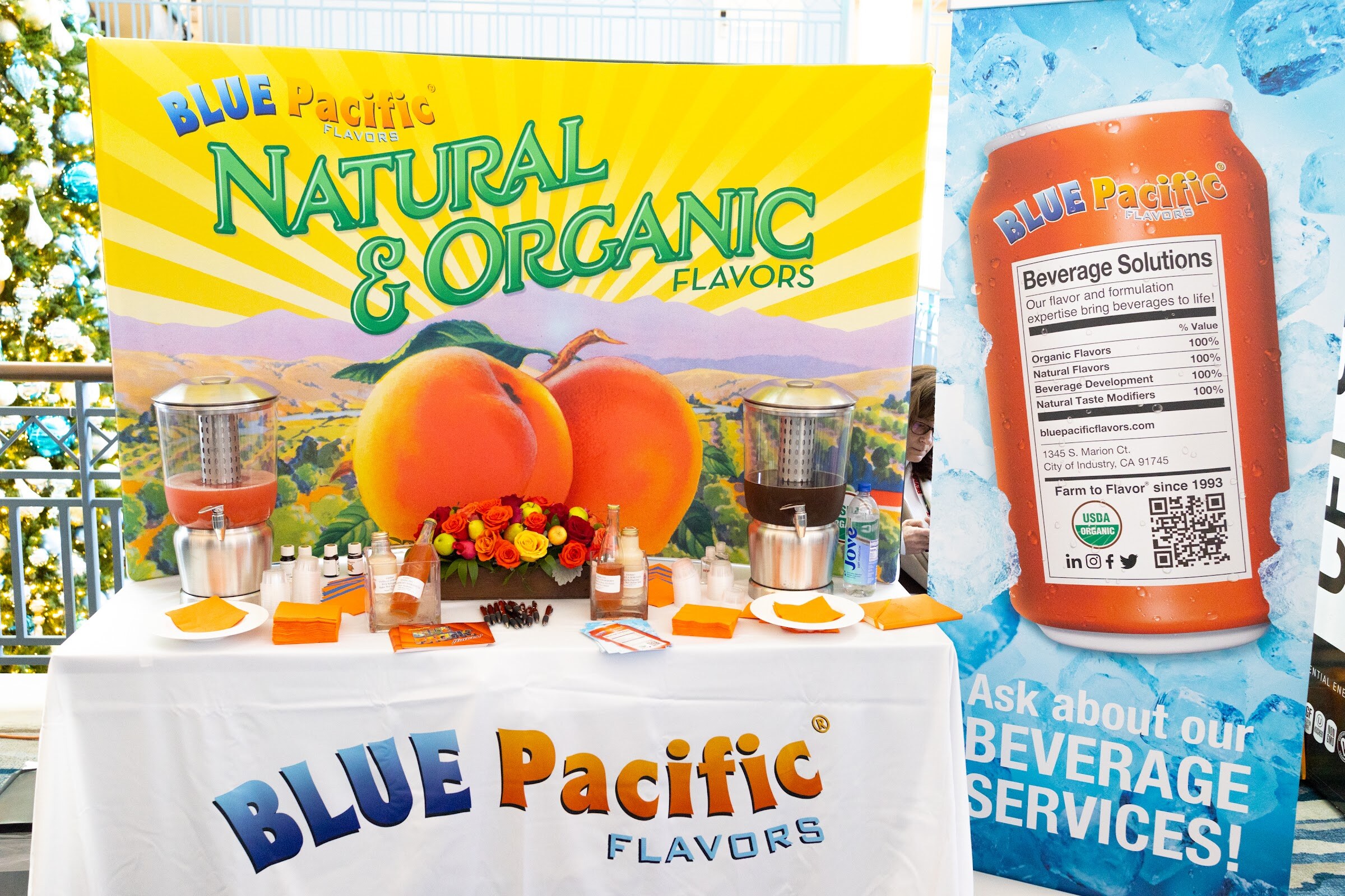 A table with a Blue Pacific Flavors cloth holds a display of peaches, flowers and sample cups in front of a yellow Natural and Organic Flavors sign and a blue banner with a can.