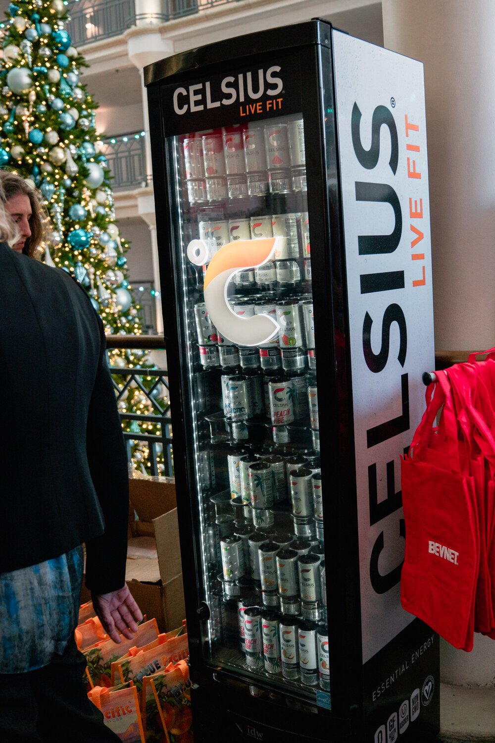 A tall lit cooler labeled Celsius Live Fit holding rows of cans next to a decorated Christmas tree.