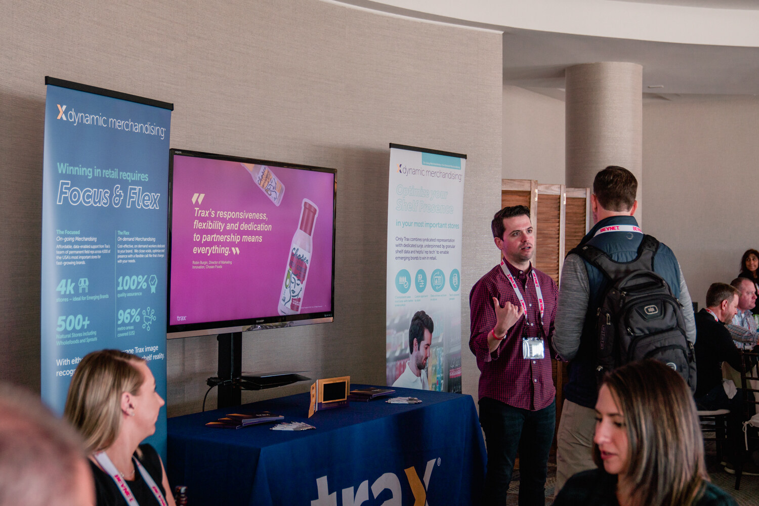 A blue-draped table with a screen and banners for Dynamic Merchandising and Trax, with attendees talking in front of them.