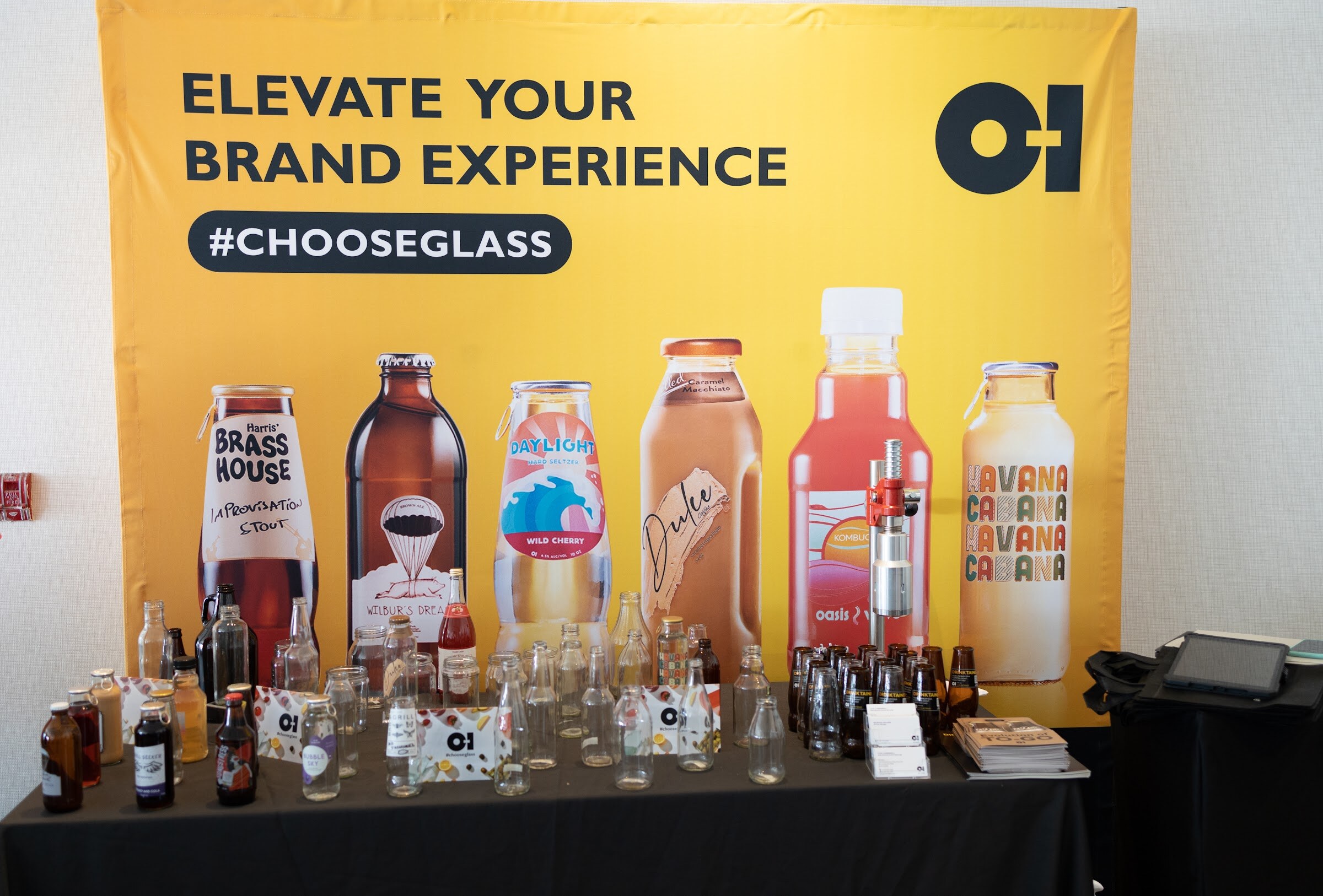 A table of glass bottles in front of a yellow banner reading Elevate your brand experience #ChooseGlass with the O-I logo.