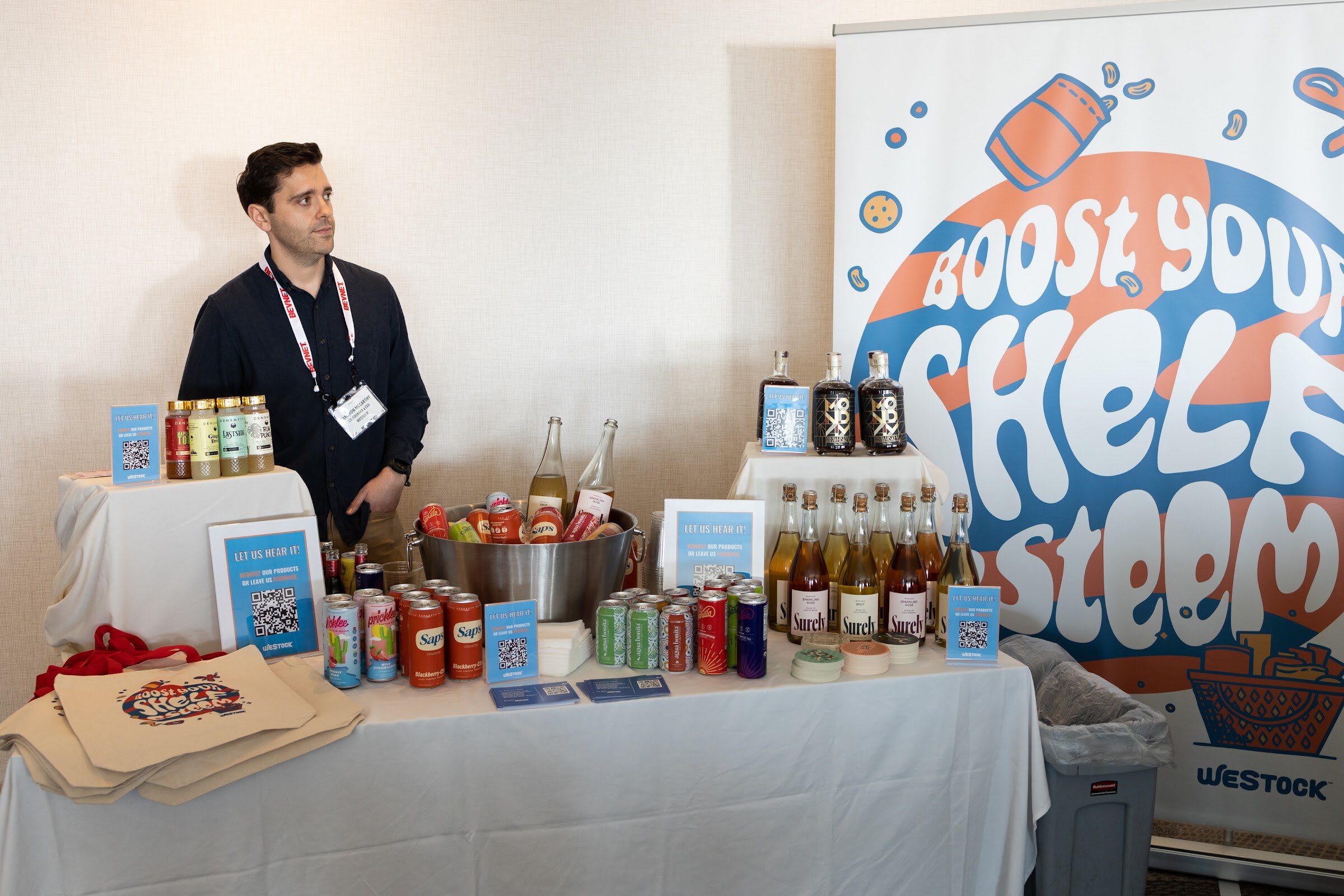 A man in a navy shirt with a BevNET lanyard stands behind a white-clothed table with cans, bottles, QR code signs and tote bags.
