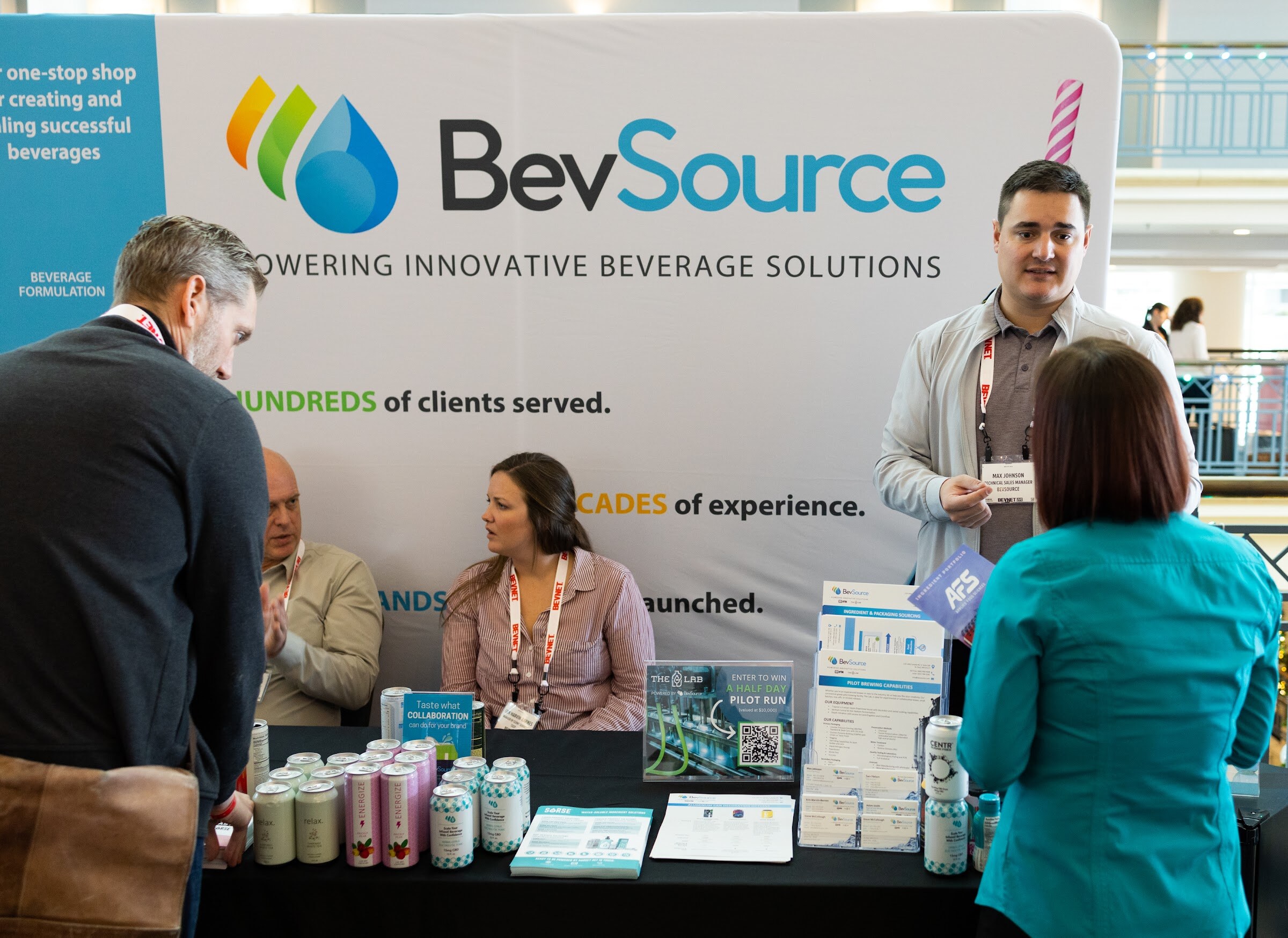 Several people at a booth with a banner reading BevSource Powering Innovative Beverage Solutions, with cans and brochures on the table.