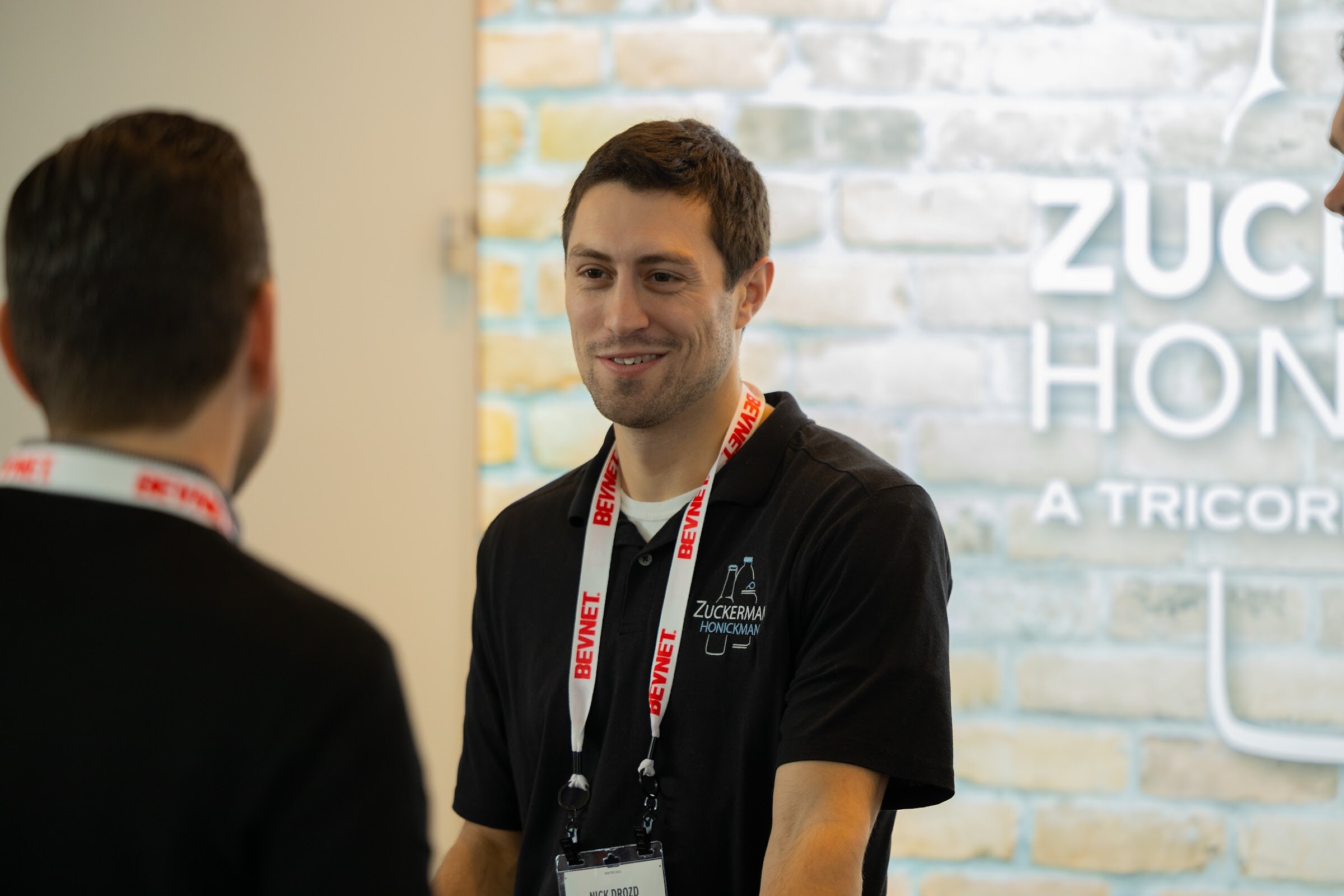 A man in a black polo with a Zuckerman Honickman logo and a BevNET lanyard smiles at someone out of frame, with a brick wall behind him.