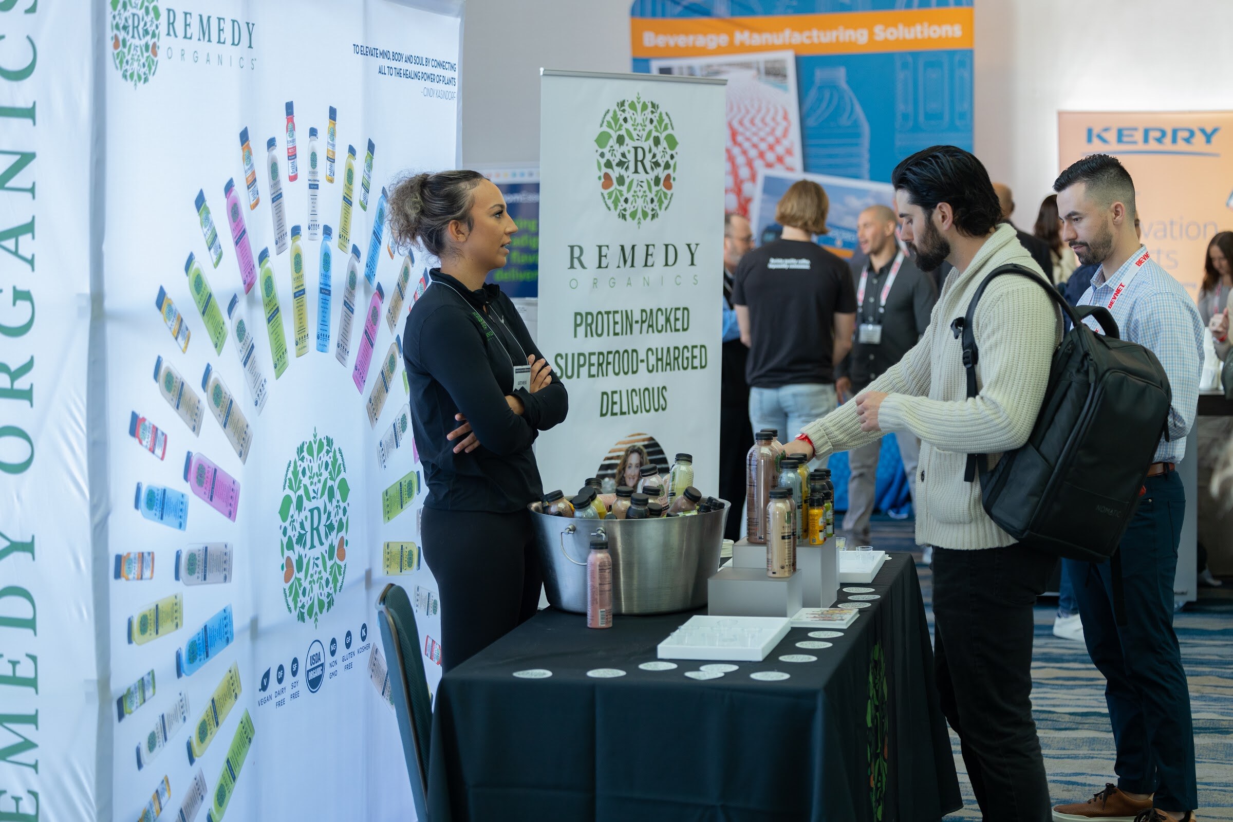 A woman in black stands with arms crossed at a table with bottles in an ice bucket in front of a Remedy Organics display.