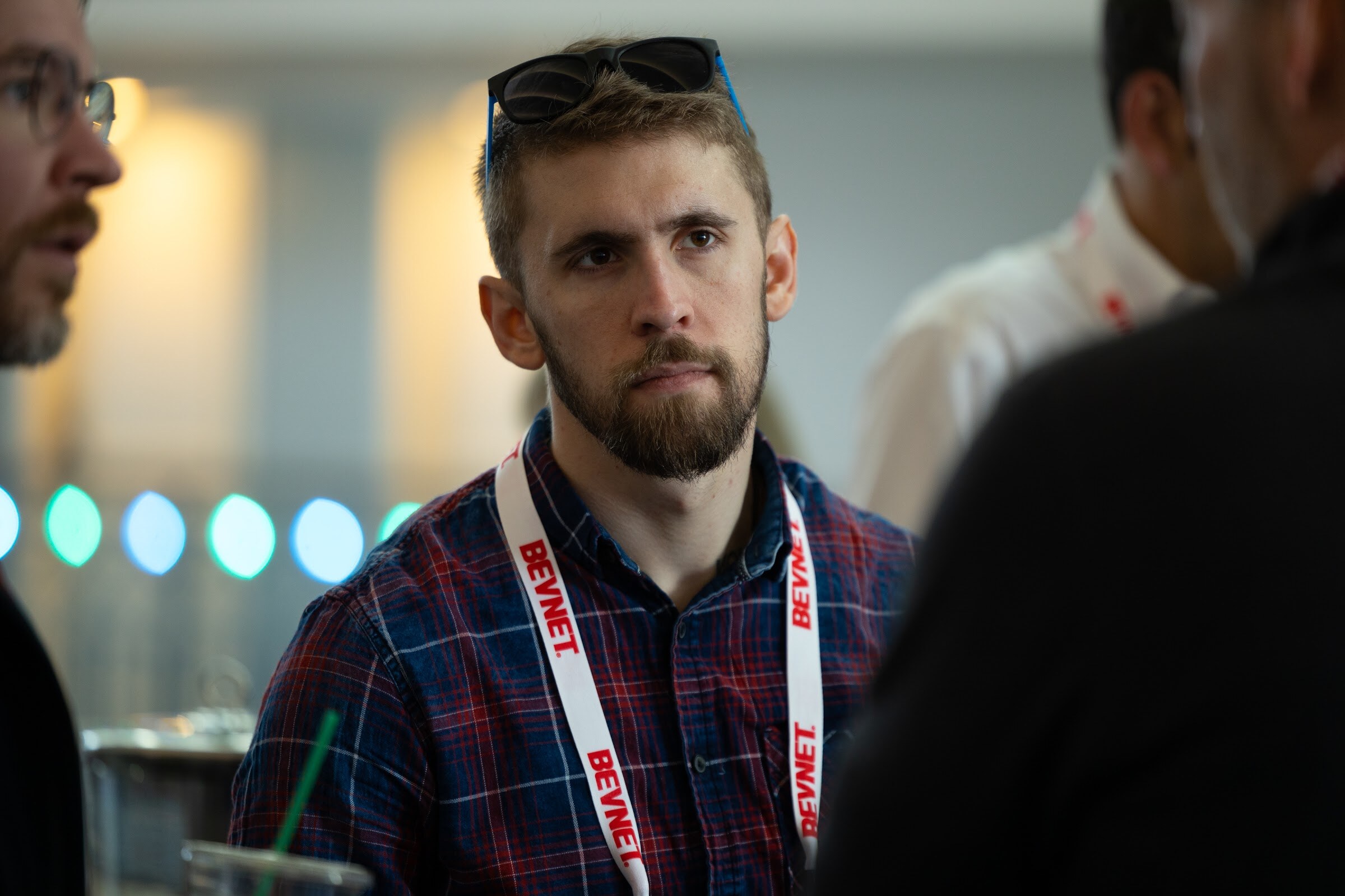 A bearded man in a red and blue plaid shirt with a BevNET lanyard looks off to one side in a hallway with lights behind him.