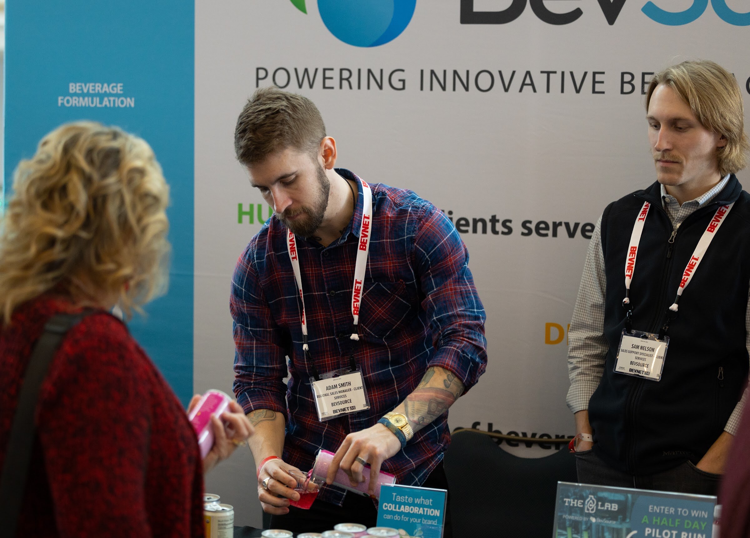 A bearded man in a plaid shirt with a BevNET lanyard pours a pink drink while a man in a vest and a woman with curly hair stand at the BevSource booth.