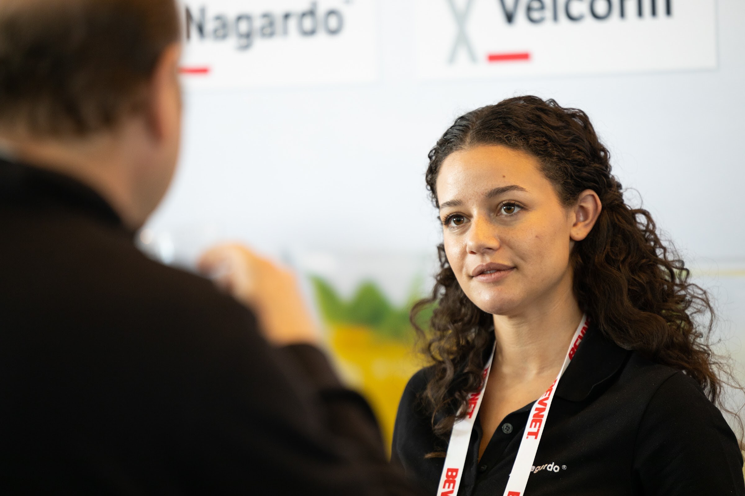 A woman with curly dark hair in a black polo and a BevNET lanyard looks at an attendee seen from behind in front of Nagardo and Velcorin signs.