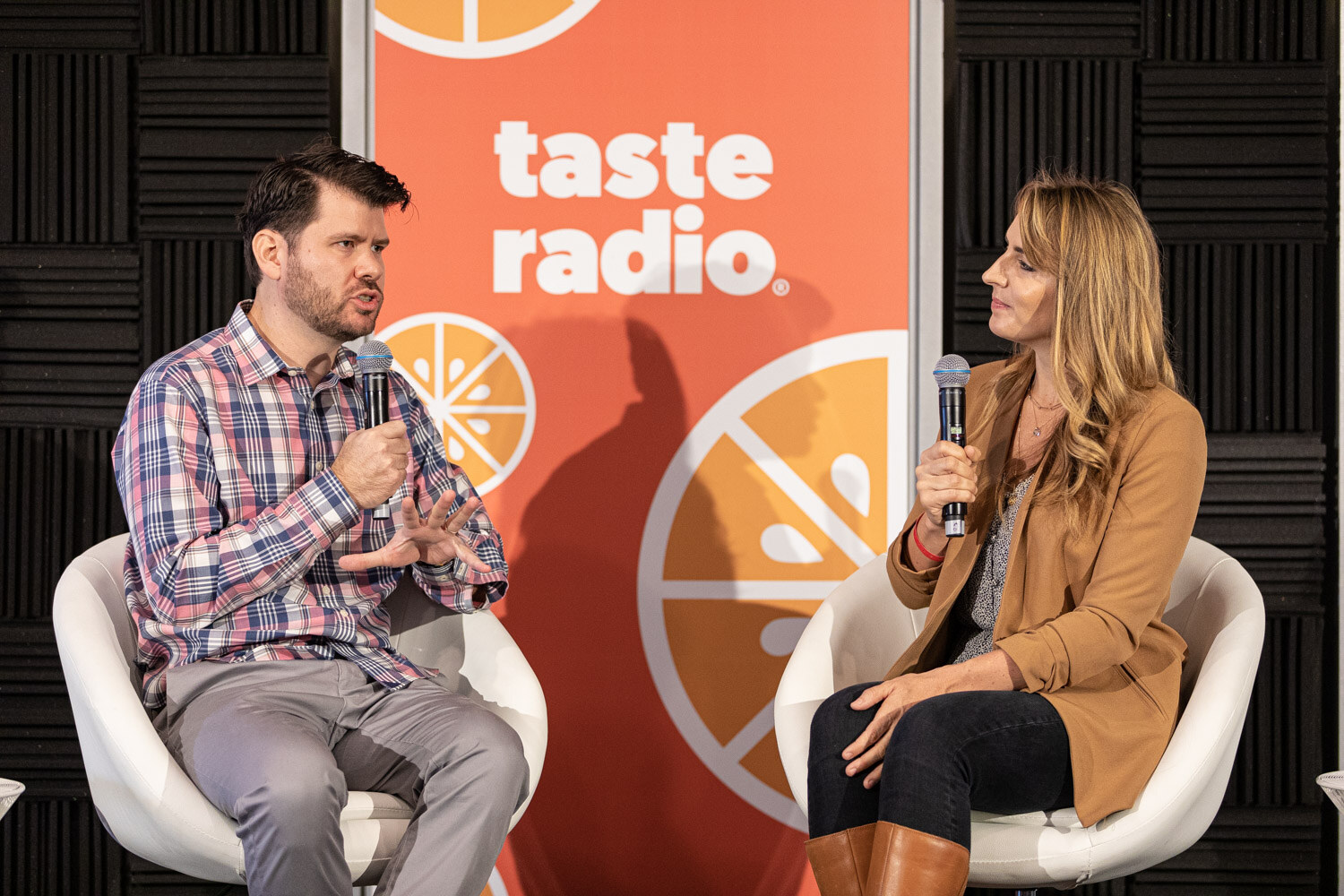 A man in a plaid shirt and a woman in a camel jacket hold microphones in white chairs in front of a Taste Radio banner.