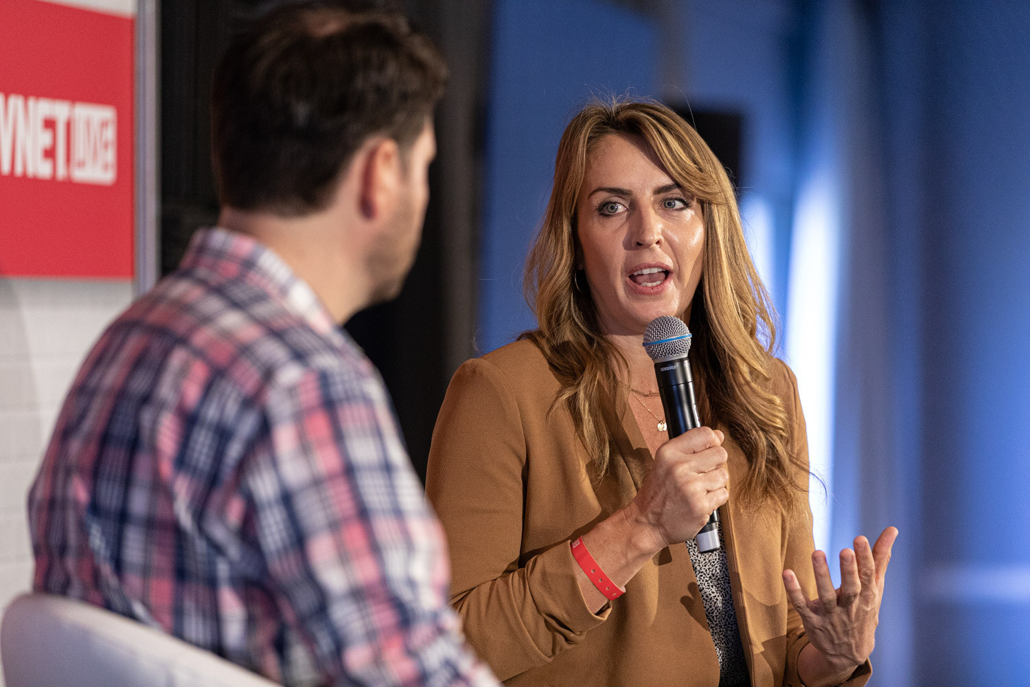A woman with long wavy hair in a camel blazer holds a microphone and gestures while a man in a plaid shirt, seen from behind, faces her.