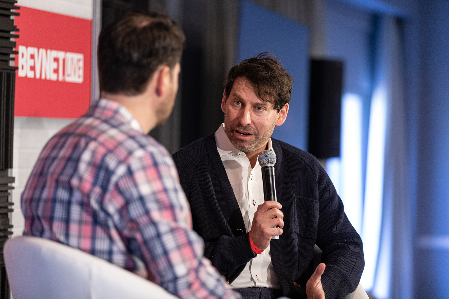 A man in a navy cardigan and white shirt holds a microphone and talks to a man in a plaid shirt seen from behind.