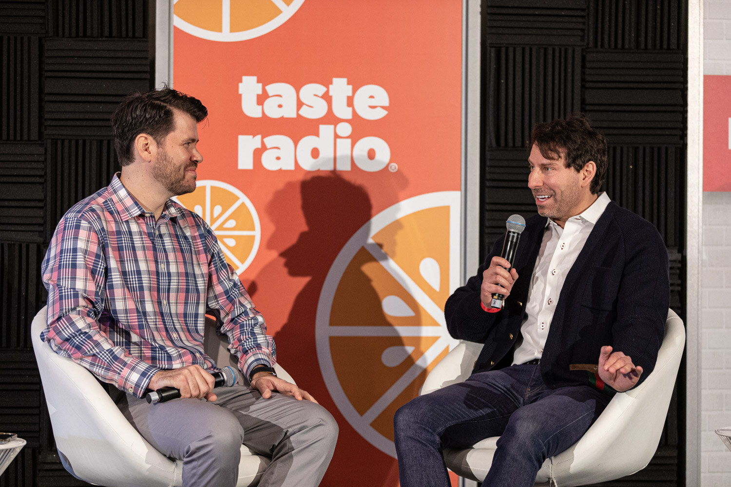A man in a plaid shirt and a smiling man in a navy cardigan sit in white chairs in front of a Taste Radio banner.