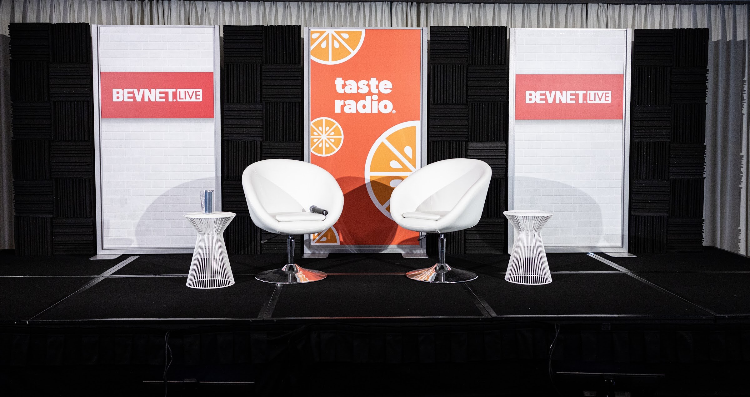 An empty stage with two white round chairs and side tables in front of a Taste Radio banner flanked by two BevNET Live signs.